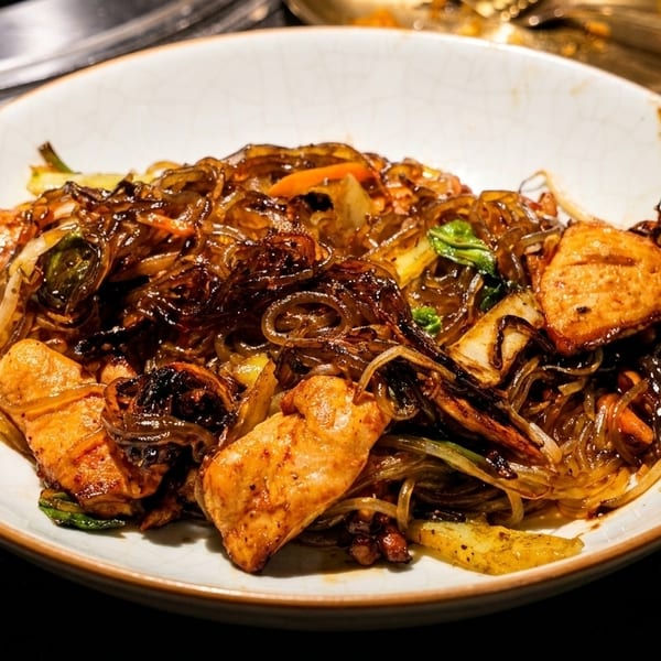 Close-up of a bowl of Korean japchae with glass noodles, vegetables, and chicken pieces.